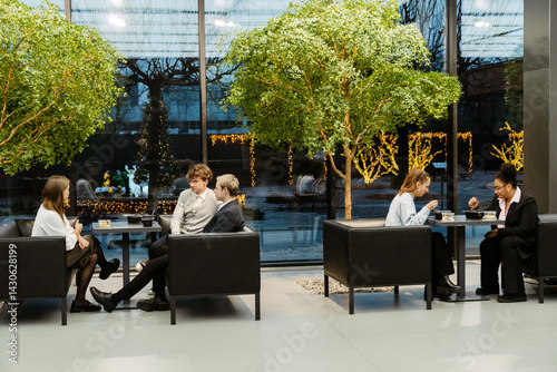 Two multiethnic formally dressed groups of people, including White and Black, sit at separate tables, talk, and have a supper break at work, in a modern office lobby with trees, and a glass wall.
