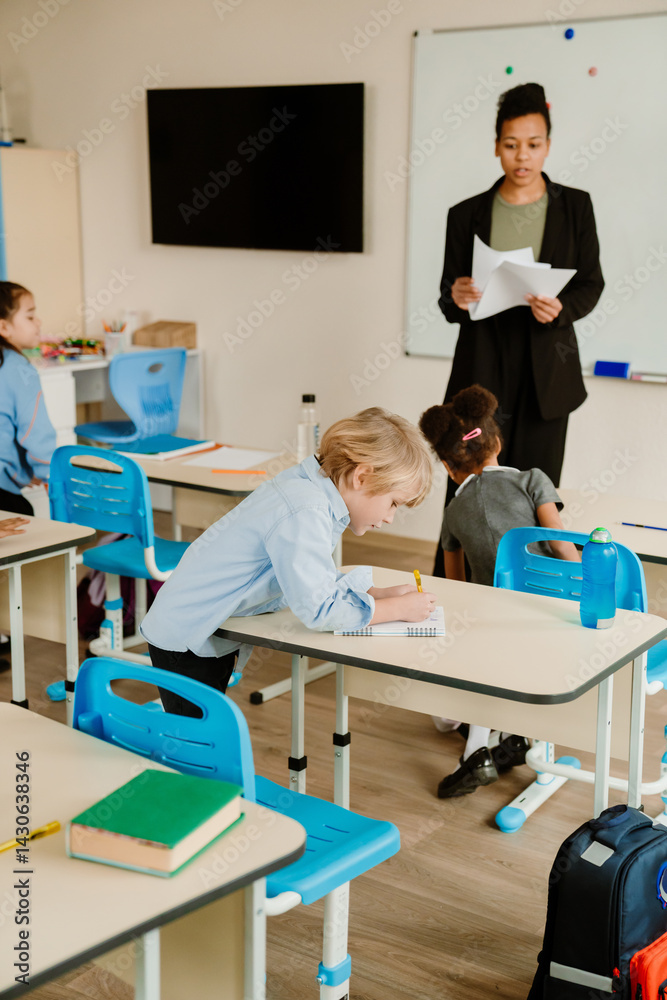 Obraz premium A classroom with an adult Latino female teacher addressing diverse little students. A young White boy writes at his desk, while other children, including a Black girl, are seated nearby.