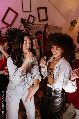 A 25-year-old brunette Asian woman wearing a denim body suit, dancing together with her mid-20s Latina female friend at a house party, singing into wireless microphones and having fun