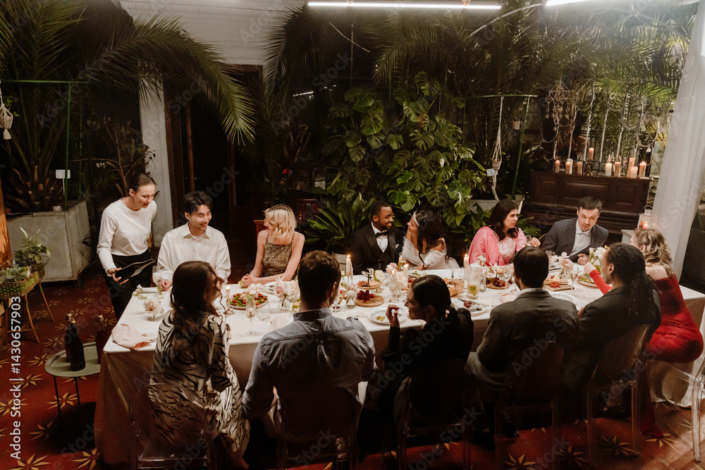 In a plant-filled wedding conservatory, a multiethnic group of guests sit talking at a long candlelit dinner table with a marrying Black couple in their 20s, while a White female server pours wine.
