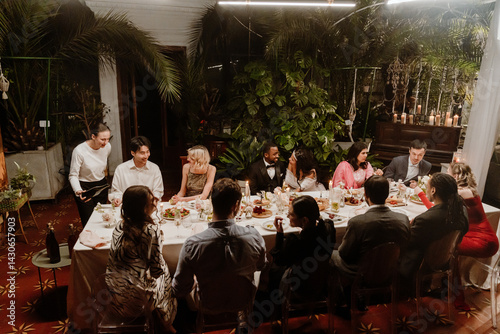 Obraz na płótnie In a plant-filled wedding conservatory, a multiethnic group of guests sit talking at a long candlelit dinner table with a marrying Black couple in their 20s, while a White female server pours wine. w salonie