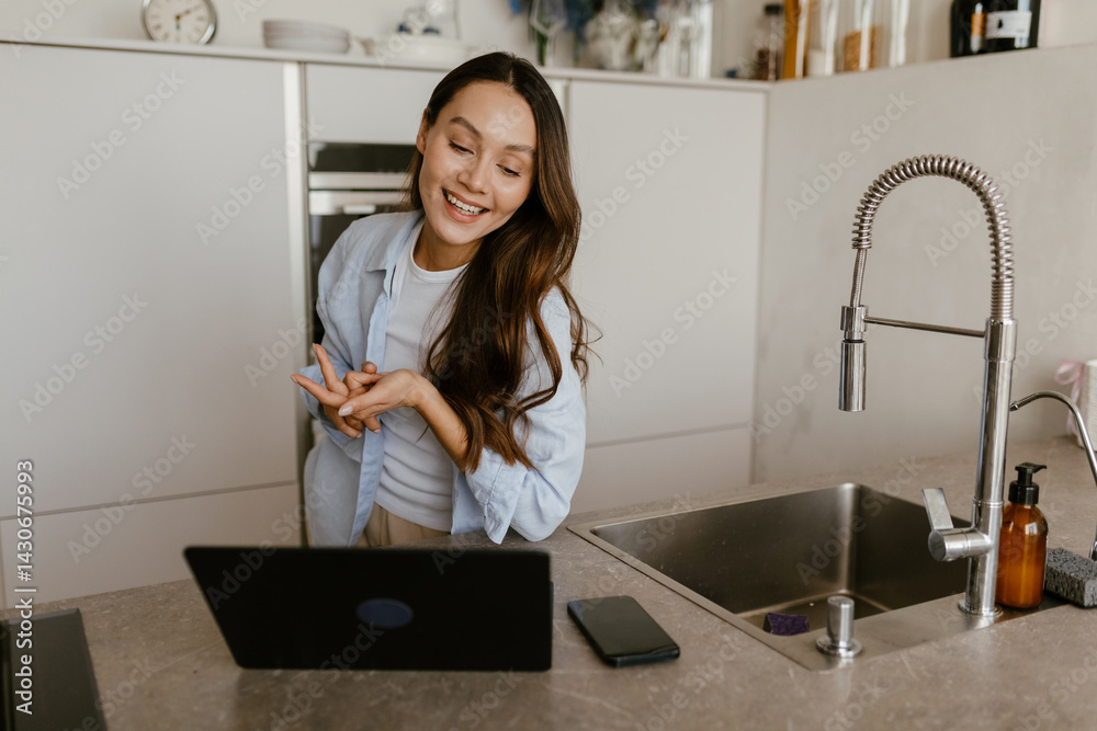 A calm European brunette woman aged 25, smiling widely while looking at her laptop as she stands next to the sink in a modern, minimalistic kitchen, talking to someone on videocall