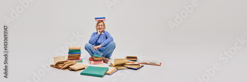 Obraz na płótnie Senior woman sitting cross-legged with one book on head, surrounded by colorful books on floor against white studio background. Concept of education for older adults, active aging w salonie