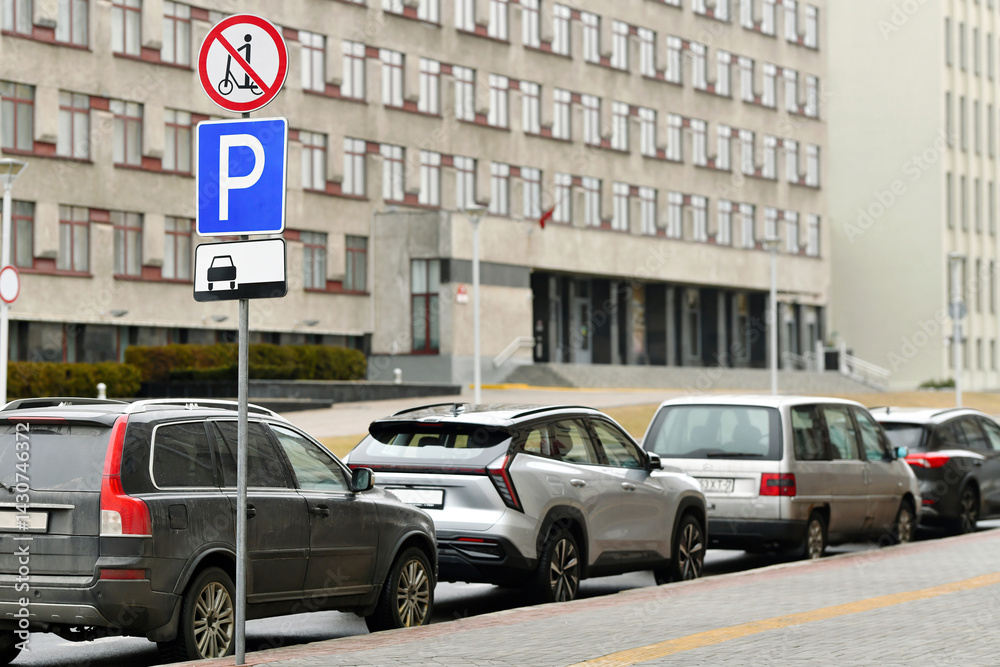 Row of parked vehicles along city sidewalk in front of government-style building with visible parking and restriction signs, no scooter zone warnings on cloudy day. Parking with multiple cars lined up