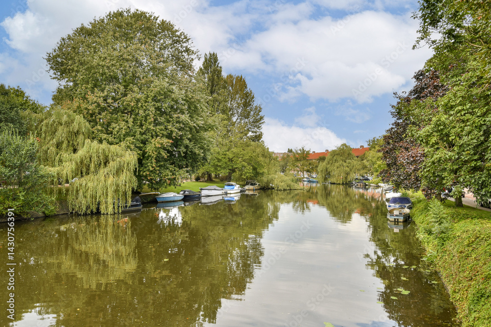 A picturesque view of a calm river bordered by lush greenery and colorful boats gently floating on the water reflecting the vibrant trees. A picturesque view of a calm river bordered by lush greenery and colorful boats gently floating on the water reflecting the vibrant trees.