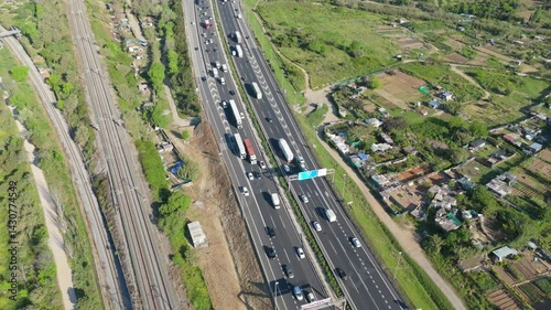 Capturing an aerial view of a highway and railway tracks weaving through a rural landscape with cultivated allotments and small houses, highlighting transportation infrastructure