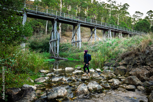 Fotografie Woman crossing the river with an old timber trestle bridge in the background, Ti
