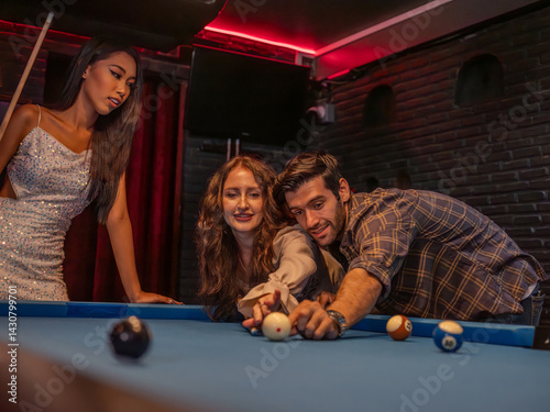 Three friends share a joyful moment at a pool table in a cozy bar. A man lines up his shot, leaning in with focus, while one woman cheerfully