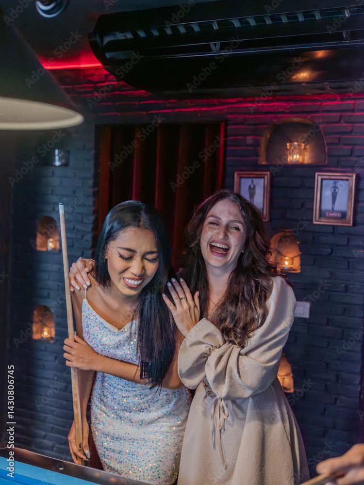 Two female friends share a moment of laughter beside a pool table in a trendy bar. One woman leans on her cue, laughing with eyes closed,