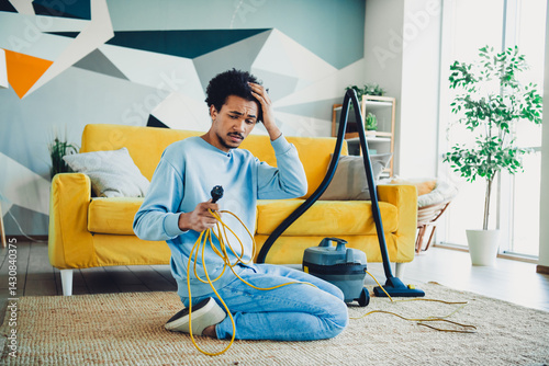Φωτογραφία Young man seated on floor struggling with tangled vacuum cord in bright modern l