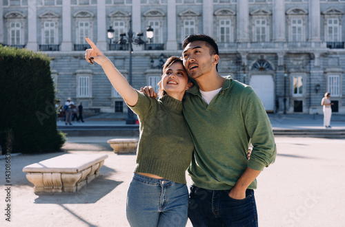 Excited couple exploring Madrid, woman pointing at landmark while man looks curiously. Joyful moment of discovery and cultural appreciation in historic urban setting