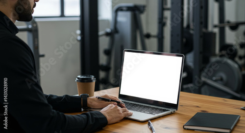 A man in a black jacket working on a laptop in a gym. He is seated at a wooden table, typing on a laptop with a blank screen, a coffee cup nearby, and gym equipment out of focus in the background.
