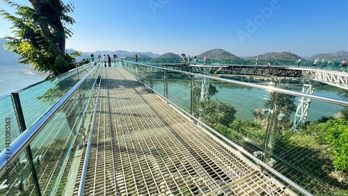 A scenic walkway with a glass railing offers stunning views of mountains and a body of water, inviting visitors to explore the natural beauty around them.