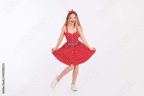 Smiling young woman dressed in a vintage red polka dot dress posing charmingly on a white background, embodying retro and pinup style