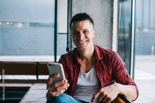 Smiling man in casual wear using cellphone sitting near window