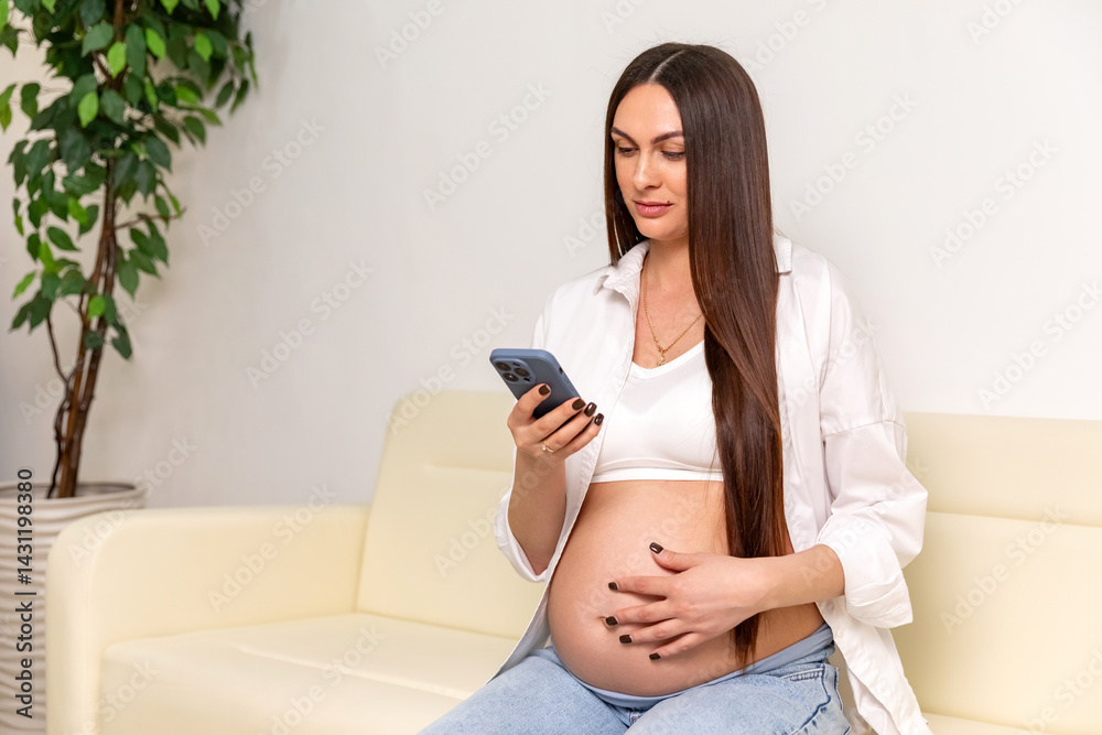 Happy pregnant woman using a smartphone and gently touching her belly while relaxing on couch at home. Happy young expectant mother. Family planning and IVF centers concept.