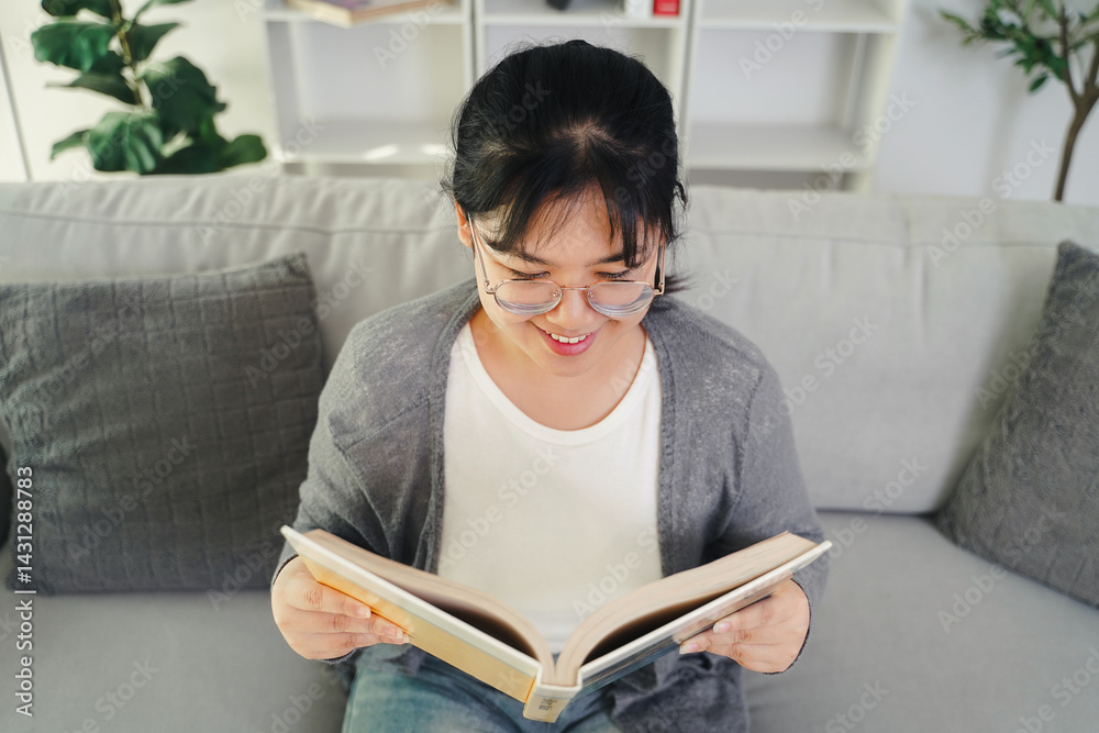 Asian young woman with glasses reading a book on the sofa in the living room happily Asian young woman with glasses reading a book on the sofa in the living room happily