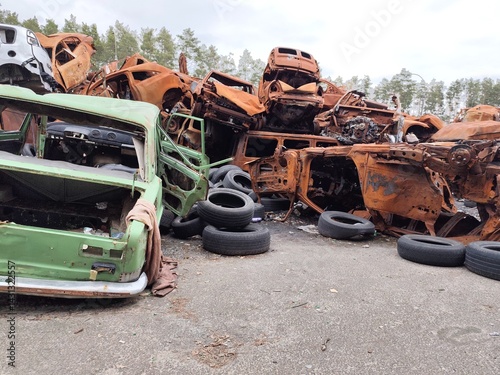 A haunting tableau of rusted, bullet-riddled civilian vehicles stacked in the streets of Irpin, Ukraine, after the 2022 invasion. Once ordinary cars lie twisted and charred beneath a silent pine