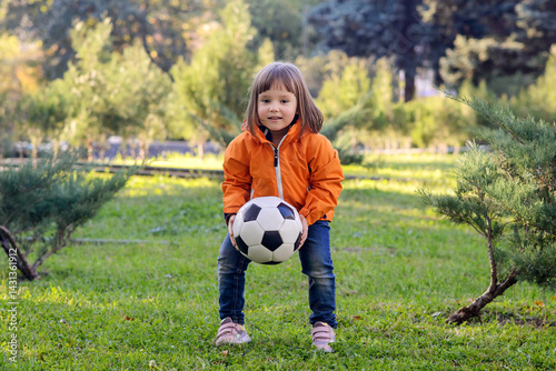 Canvas Print Little girl playing football outdoors: smiling toddler child in orange jacket holding soccer ball on green lawn in impromptu goal posts in park