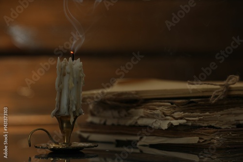 Fotografi Smoldering candle in candlestick and old books on mirror surface, closeup