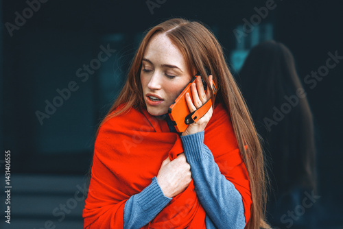 Woman wrapped in a red shawl making a phone call outdoors during a cool day with a reflective building background