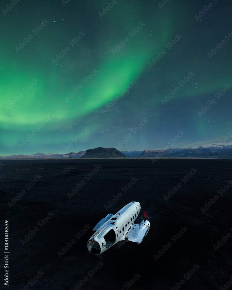 DC-3 wreck on Solheimasandur’s black sand beach beneath soft green Northern Lights, with a red-clad figure standing for contrast and scale in the vast night landscape.