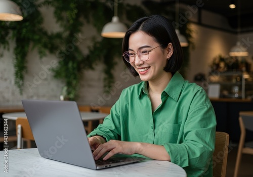 Smiling woman works on laptop at cafe table, green shirt, glasses