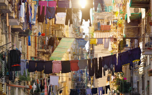 Street in Naples, Italy, with clothes hanging on lines between old buildings. The vibrant laundry contrasts with weathered walls, balconies, and sunlit facades, authentic southern Italian urban scene