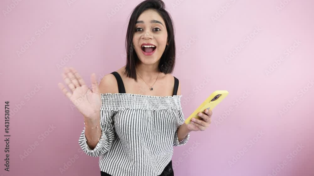South Asian young woman holding smartphone, looking at camera and saying hi with waving hand gesture