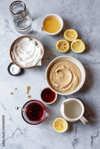 An assortment of ingredients on a marble surface including coconut cream, lemon juice, maple syrup, tahini, and a glass of water.