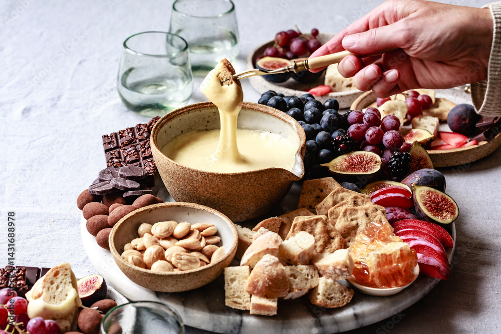 An assortment of fruits, nuts, and bread surrounds a bowl of cheese fondue, with a hand dipping a piece of bread into it. An assortment of fruits, nuts, and bread surrounds a bowl of cheese fondue, with a hand dipping a piece of bread into it.