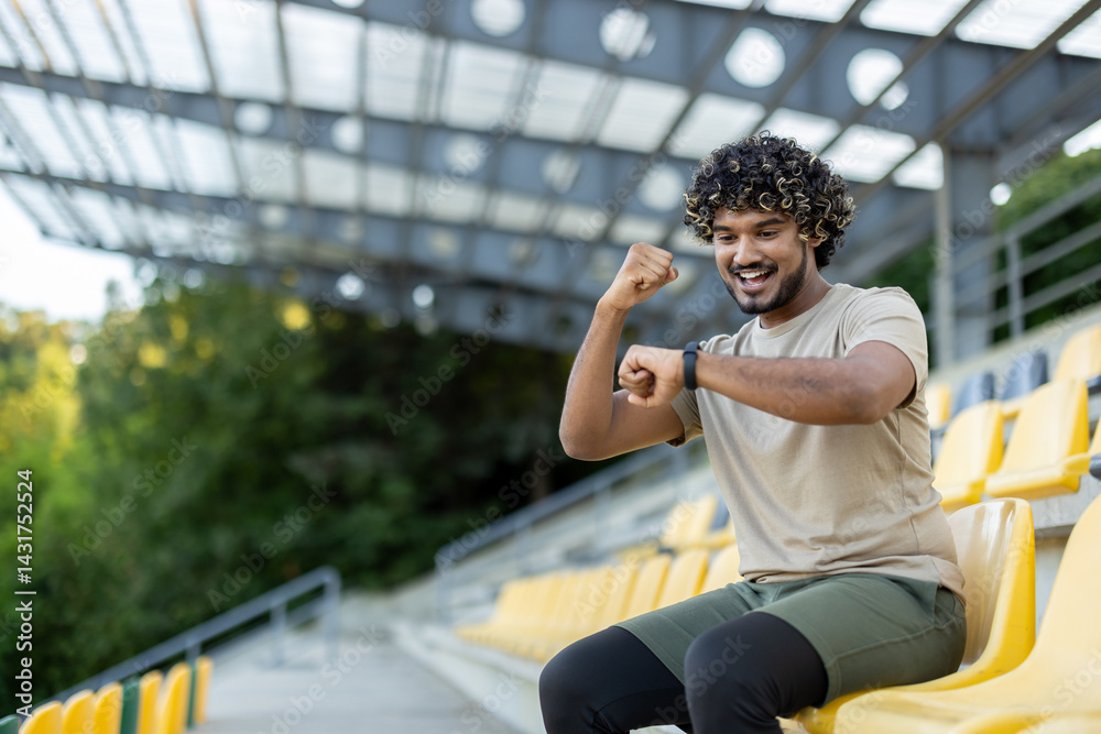 Young athlete celebrates successful training results, resting while sitting. Man checks data on smart watch fitness bracelet, smiles contentedly, holds hand up.