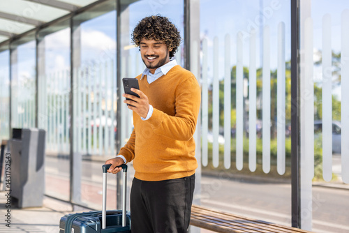 A smiling man looks at his phone while waiting at a bus stop, luggage in hand. He is dressed in a sweater and a collared shirt