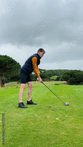 Man prepares and swings at a golf ball on a well-kept course under an overcast sky, dressed in sporty gear with focused posture and motion.
