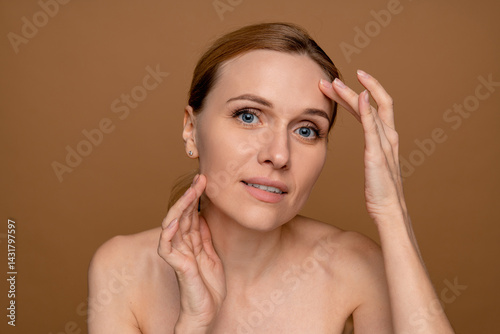 Aged beauty concept. Portrait of beautiful middle age woman touching silky skin on her face, smiling at camera over light studio brown beige wall background. Charming lady pampering herself