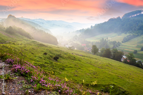 Obraz na płótnie abranka village in the valley at foggy sunrise. countryside landscape with red clouds above forested hills and rural fields. beautiful scenery of transcarpathia, ukraine in summer. grassy meadow w salonie