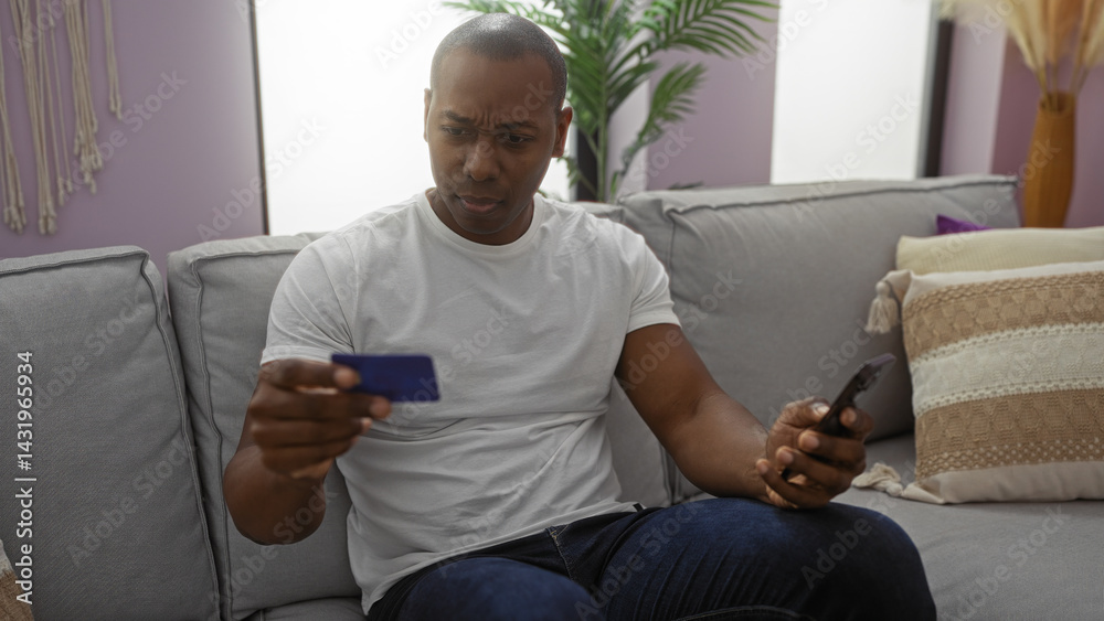 Young man inside living room looks at credit card with a concerned expression holding phone on couch in modern apartment setting with indoor plants and cozy decor around.