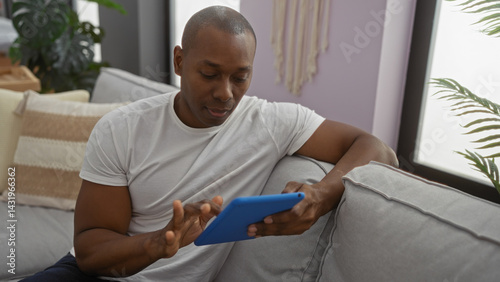 Young man using tablet at home sitting on a comfortable couch indoors with natural lighting and modern decor surrounding him in a relaxed living room setting.