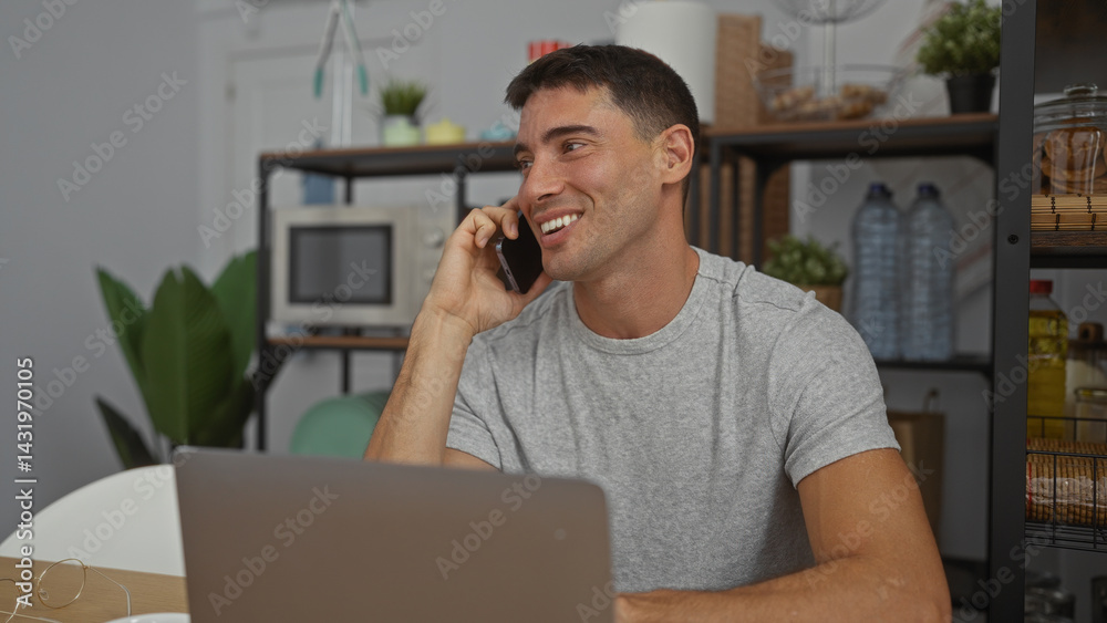 Young man in office talking on phone with smile during work, laptop in front, indoor setting showing modern workplace decor with plants and shelves.