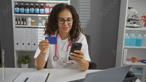 Obraz na płótnie Mature woman in a clinic wearing a stethoscope holds a credit card and smartphone while working in a medical office setting with shelves of supplies and an organized desk environment. w salonie