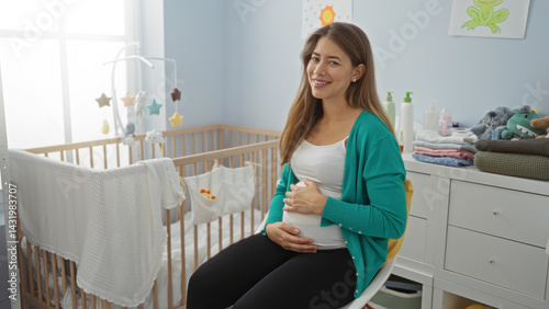Pregnant woman smiling while sitting in a cozy nursery room, surrounded by baby items, cradling her belly with happiness and anticipation in a bright home environment.