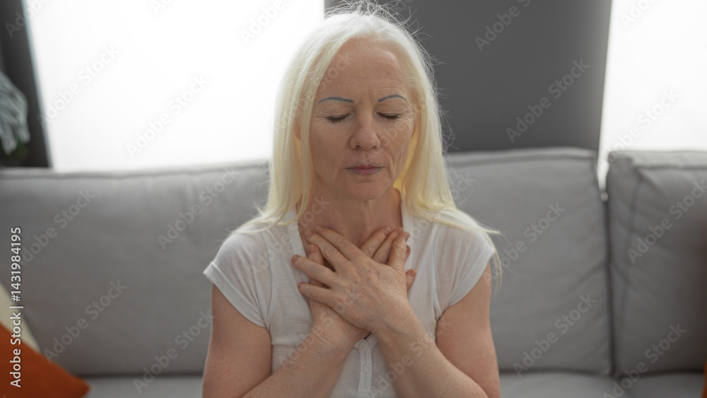 Woman meditating in a bright living room, displaying tranquility and calm, with her hands gently placed on her chest, reflecting a serene atmosphere of relaxation and peace at home.