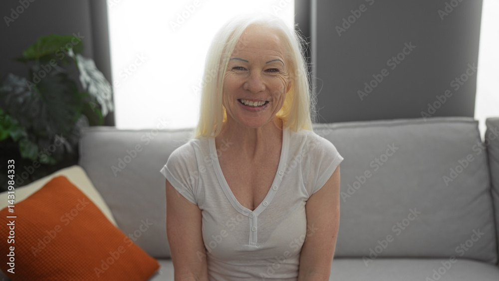 Woman smiling indoors in a cozy living room with a white shirt and blonde hair, sitting on a couch with colorful pillows, capturing a warm and inviting atmosphere.