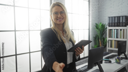 Obraz na płótnie Woman smiling in office extends handshake holding tablet in modern workplace with computers, glass windows, and bookshelves, highlighting professional and friendly interaction. w salonie