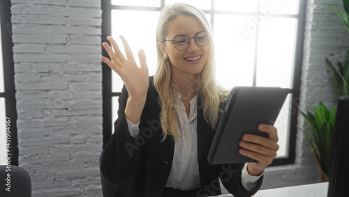 Obraz na płótnie Woman smiling in office while video chatting on tablet, wearing glasses, with bright interior, brick walls and indoor plants creating a professional workspace environment. w salonie
