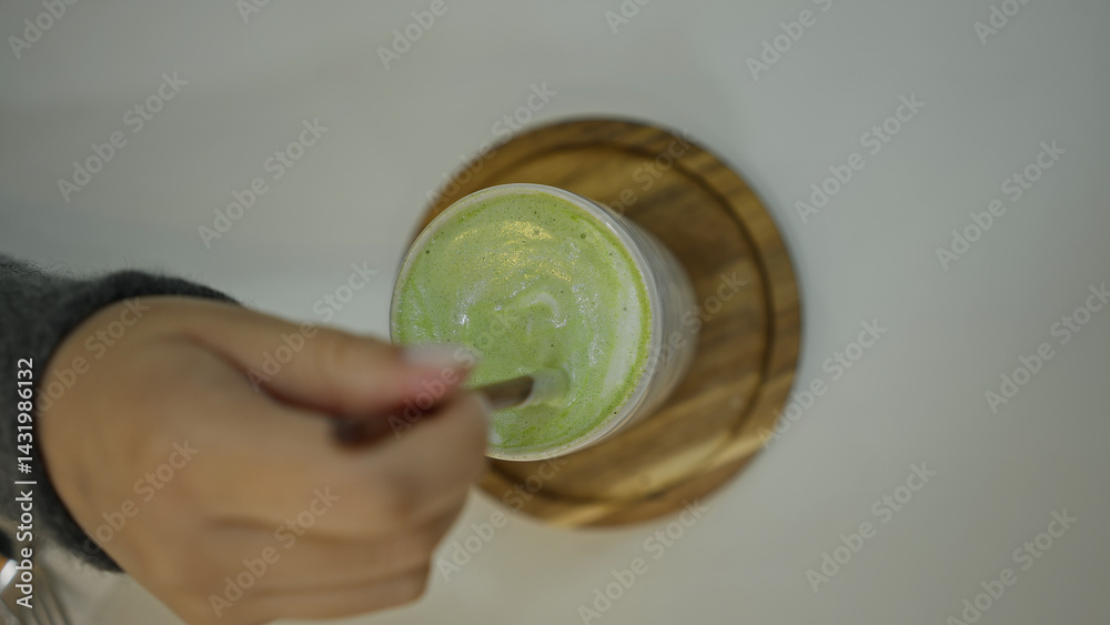Woman stirring matcha latte with spoon inside cozy coffee shop creating soothing drink atmosphere with wooden coaster adding rustic elegance to the setting.