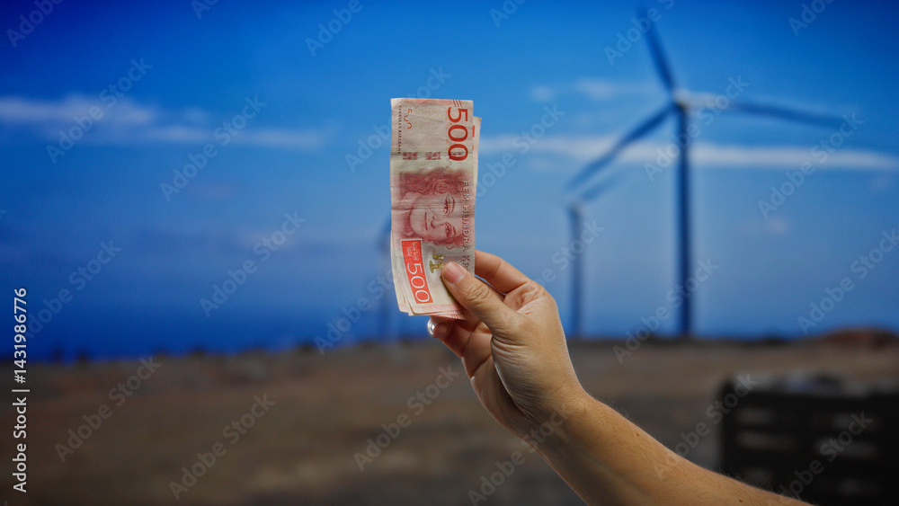 Hand holding swedish krona in front of windmills in a field, symbolizing investment in sustainable energy and eco-friendly financial growth outdoors.