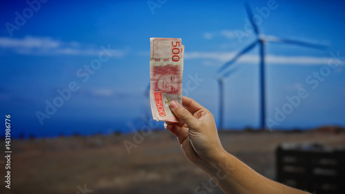 Obraz na płótnie Hand holding swedish krona in front of windmills in a field, symbolizing investment in sustainable energy and eco-friendly financial growth outdoors. w salonie