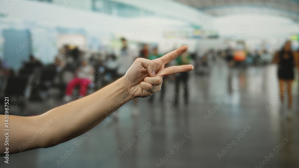 Man gesturing peace sign indoors at busy airport with flight passengers in background, showcasing travel atmosphere and human connection.