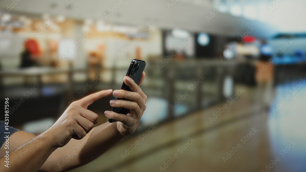 Man holding smartphone in indoor shopping mall with blurred background, focusing on touchscreen, reflecting modern retail technology and communication.
